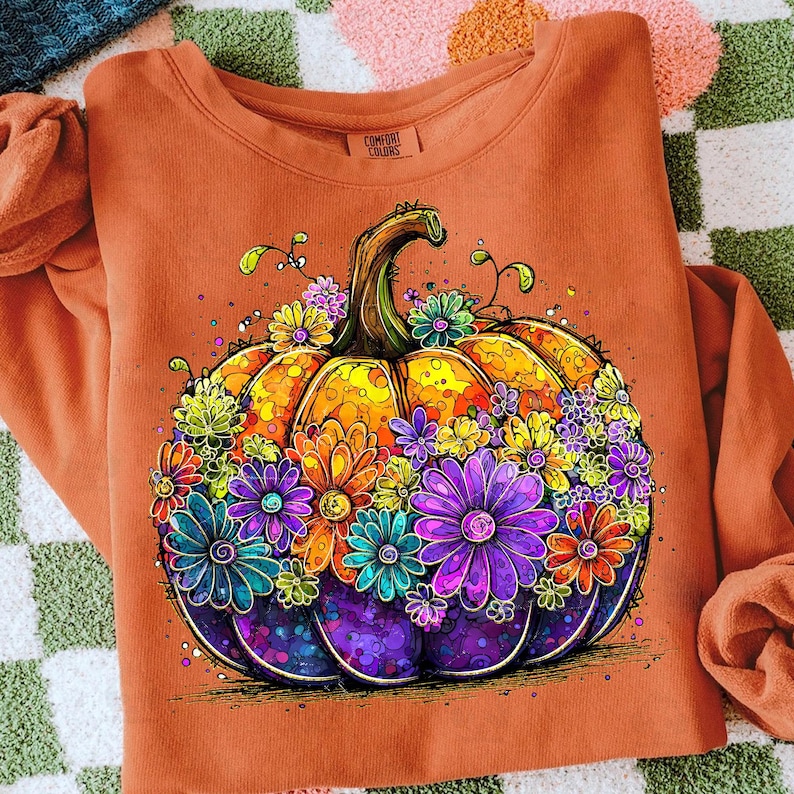 May include: An orange sweatshirt featuring a colorful, detailed illustration of a pumpkin decorated with vibrant flowers. The pumpkin has a yellow and orange body with a green stem, and is surrounded by purple, blue, and orange flowers.