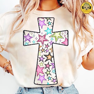 Puede incluir: Una camiseta color crema con un diseño de cruz grande lleno de estrellas de colores. Las estrellas son en tonos de rosa, morado, azul, amarillo y naranja, con contornos negros. La cruz tiene un borde negro.