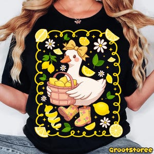 May include: Black t-shirt featuring a cartoon goose with a yellow bow, holding a basket of lemons. The design includes lemons, daisies, and lemon peel accents, with yellow rain boots. The shirt has the text "GrootStoree" at the bottom.