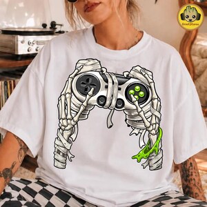 Op de afbeelding: Een wit t-shirt met een afbeelding van twee in verband gewikkelde skeletthanden die een videogamecontroller vasthouden. De controller is grijs en zwart met groene knoppen. Een beetje groen slijm druipt van de rechterkant van de skeletthand.
