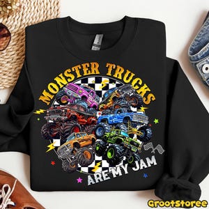 K&ouml;nnte beinhalten: Schwarzes Sweatshirt mit einem farbenfrohen Monster-Truck-Motiv. Das Design zeigt verschiedene Trucks in Lila, Orange, Blau und Gr&uuml;n, mit dem Text "MONSTER TRUCKS ARE MY JAM" in kreisf&ouml;rmiger Anordnung.