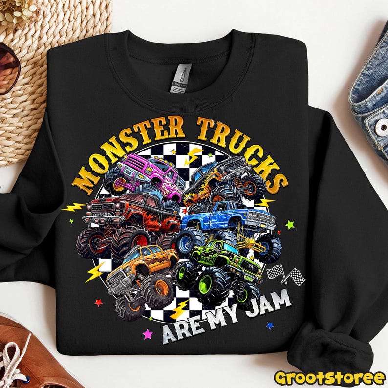 K&ouml;nnte beinhalten: Schwarzes Sweatshirt mit einem farbenfrohen Monster-Truck-Motiv. Das Design zeigt verschiedene Trucks in Lila, Orange, Blau und Gr&uuml;n, mit dem Text "MONSTER TRUCKS ARE MY JAM" in kreisf&ouml;rmiger Anordnung.