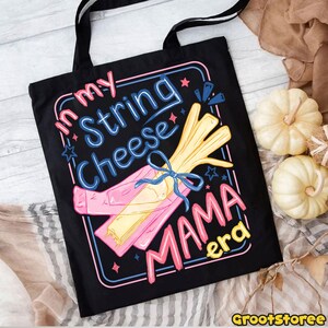 May include: Black tote bag with the text "In my String Cheese Mama Era" in pink, blue, and yellow. The design features a string cheese graphic with a blue bow. The bag has black handles and is likely made of canvas.