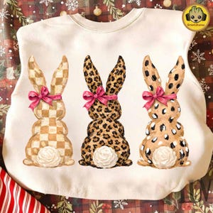 Puede incluir: Una sudadera blanca con tres figuras decorativas de conejos. Los conejos tienen diferentes patrones: a cuadros, estampado de leopardo y moteado. Cada conejo tiene un lazo rosa y una cola blanca y esponjosa. El logotipo de GrootStore está en la esquina superior derecha.
