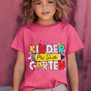 May include: A pink t-shirt featuring a colourful graphic that reads "Kinder Mrs. Smith Garten". The graphic is made up of letters with various patterns and colours, and includes a pencil with a yellow eraser.
