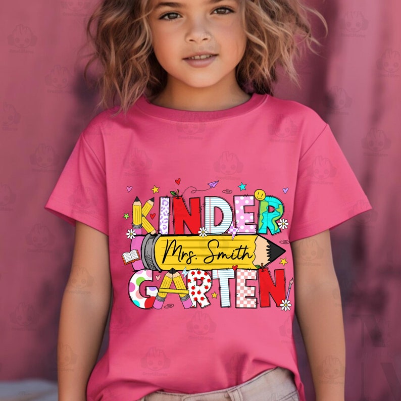 May include: A pink t-shirt featuring a colourful graphic that reads "Kinder Mrs. Smith Garten". The graphic is made up of letters with various patterns and colours, and includes a pencil with a yellow eraser.