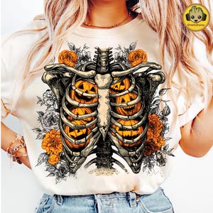 May include: Cream-colored t-shirt featuring a detailed illustration of a rib cage filled with carved jack-o'-lanterns. The design includes orange and white floral accents, creating a spooky yet artistic Halloween theme.