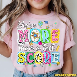 May include: Pink short-sleeved t-shirt with the text "You're MORE than a test SCORE" in colorful, block letters. The design includes school-themed graphics like a globe, apple, and stars. The shirt is worn by a child.
