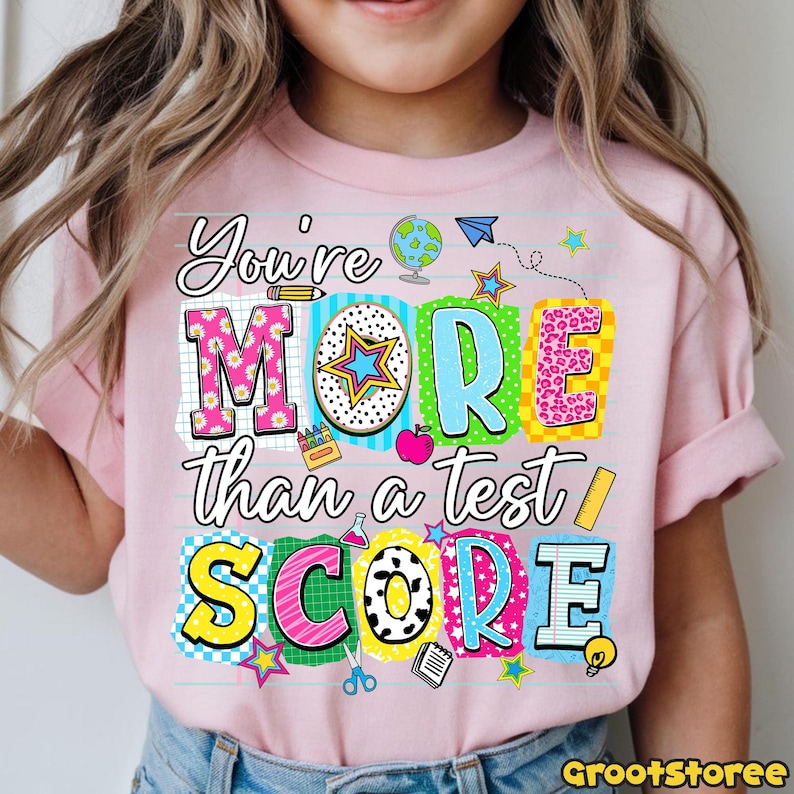 May include: Pink short-sleeved t-shirt with the text "You're MORE than a test SCORE" in colorful, block letters. The design includes school-themed graphics like a globe, apple, and stars. The shirt is worn by a child.