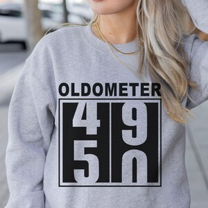 May include: A light gray sweatshirt with the word "OLDOMETER" above a black rectangle containing the numbers "49" and "50". The sweatshirt is a casual style.