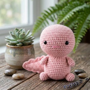 Könnte beinhalten: Ein hellrosa gehäkeltes Axolotl-Plüschtier mit einem großen, runden Kopf, kleinen, stummeligen Armen und Beinen, einem breiten Schwanz und zwei kleinen schwarzen Perlenaugen. Die Figur hat einen einfachen, horizontal aufgestickten rosa Mund.