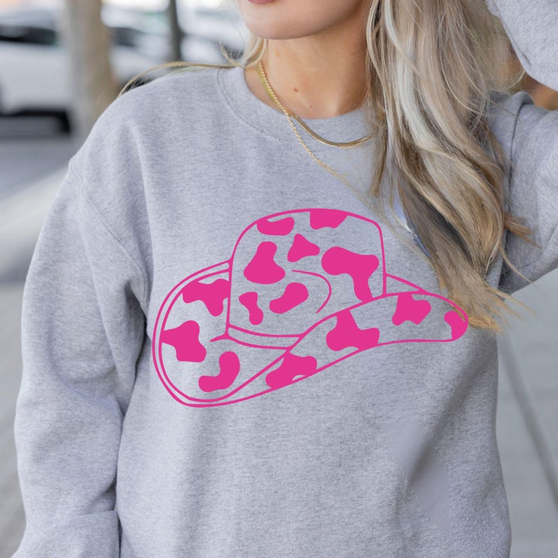 May include: A pale grey sweatshirt with a pink cowboy hat design. The hat has pink spots. The sweatshirt has a crew neck and long sleeves. The style is casual and comfortable, suitable for everyday wear.