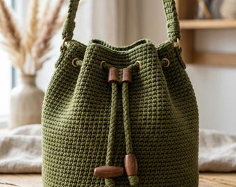 Puede incluir: Bolso de cubo de ganchillo verde oliva con cierre de cordón y presillas de madera. El bolso tiene una correa trenzada y anillos metálicos dorados. El bolso está sobre una superficie de madera.