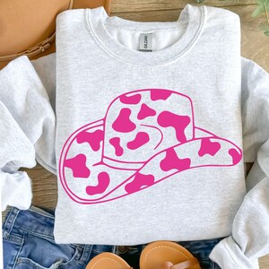 May include: A light grey sweatshirt with a large pink cowboy hat graphic featuring a cow print design. The hat is centered on the front. The sweatshirt is made of a soft material.