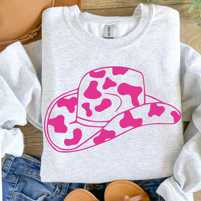 May include: A light grey sweatshirt with a large pink cowboy hat graphic featuring a cow print design. The hat is centered on the front. The sweatshirt is made of a soft material.