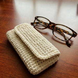 Könnte beinhalten: Ein cremefarbenes, gehäkeltes Brillenetui mit Klappenverschluss. Eine Brille mit Schildpattmuster liegt neben dem Etui auf einer Holzoberfläche. Das Etui ist handgefertigt und scheint aus Baumwollgarn zu bestehen.