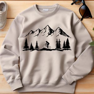 May include: A beige sweatshirt featuring a black mountain range design with a cyclist riding a bicycle. The design also includes black pine trees. The sweatshirt is made of a soft material and has a crew neck.