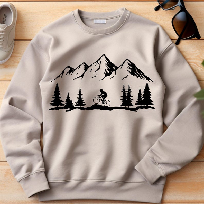 May include: A beige sweatshirt featuring a black mountain range design with a cyclist riding a bicycle. The design also includes black pine trees. The sweatshirt is made of a soft material and has a crew neck.