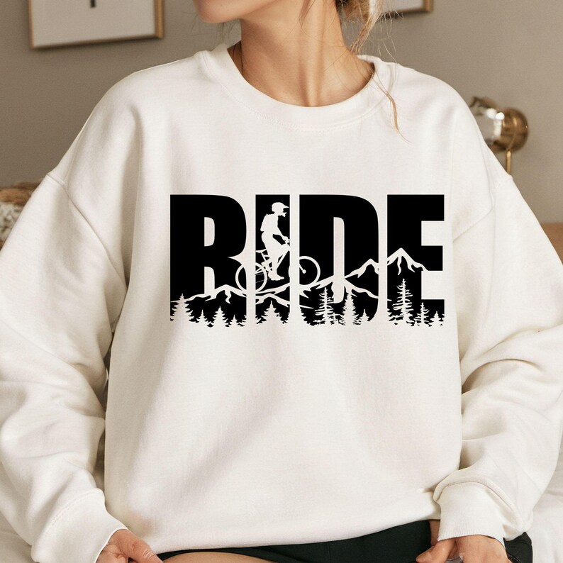 May include: Cream-colored sweatshirt with the word "RIDE" in large black block letters. The "I" in "RIDE" features a silhouette of a person riding a mountain bike, with a mountain and forest scene below.