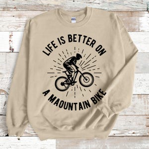 May include: A tan sweatshirt with a black graphic of a mountain biker and the text "LIFE IS BETTER ON A MOUNTAIN BIKE". The design features a cyclist on a mountain bike with a sunburst effect.