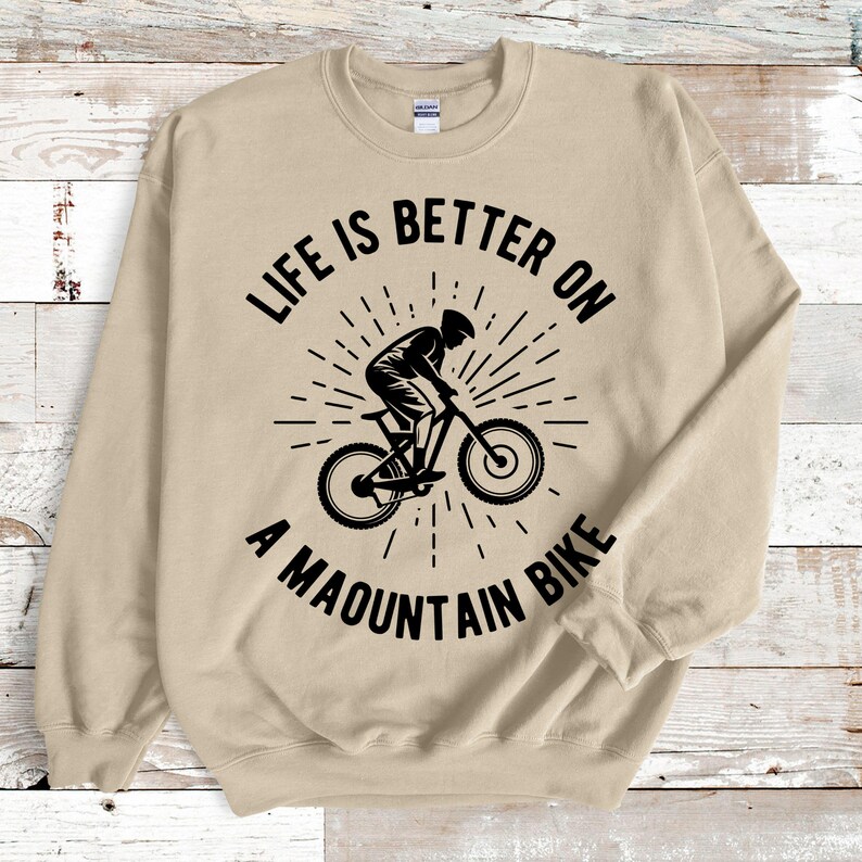May include: A tan sweatshirt with a black graphic of a mountain biker and the text "LIFE IS BETTER ON A MOUNTAIN BIKE". The design features a cyclist on a mountain bike with a sunburst effect.