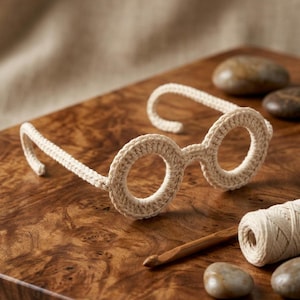 May include: A pair of beige crochet glasses featuring circular frames and thin temples. The glasses are crafted from light-colored yarn with a visible stitch pattern, showcasing a rounded design for the lenses and curved ends on the temple arms.