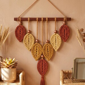 Puede incluir: Un tapiz de pared con siete adornos de ganchillo en forma de hoja en tonos óxido, mostaza y dorado. Las hojas cuelgan de una varilla de madera oscura mediante cordones anudados, decoradas con cuentas de madera y dispuestas en forma de V.