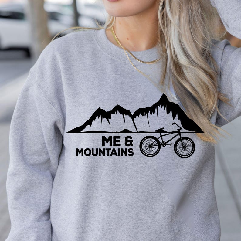 May include: A light gray sweatshirt with a black graphic of mountains, a bicycle, and the text "ME & MOUNTAINS". The sweatshirt is a crew neck style and appears to be made of a soft material.