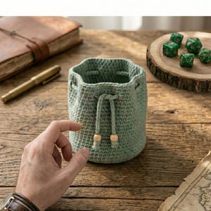 Op de afbeelding: Een saliegroene gehaakte dobbelsteenzak met een trekkoordsluiting met twee lichte houten kralen aan de uiteinden. De zak heeft een cilindrische vorm met een gestructureerd steekpatroon. De tekst CROCHET DICE BAG en INSTANT DOWNLOAD is zichtbaar.