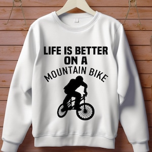 May include: White sweatshirt with the text "LIFE IS BETTER ON A MOUNTAIN BIKE" in black. A black silhouette of a person riding a mountain bike is below the text. The sweatshirt has a crew neck and long sleeves.