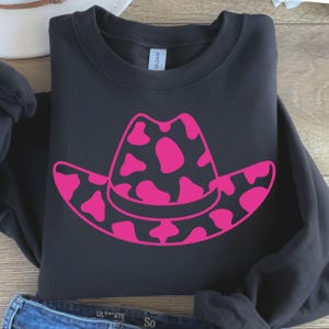 May include: Black sweatshirt with a large pink cowboy hat graphic, featuring a cow print design. The hat is centered on the front of the folded garment. The sweatshirt is made of a soft material.
