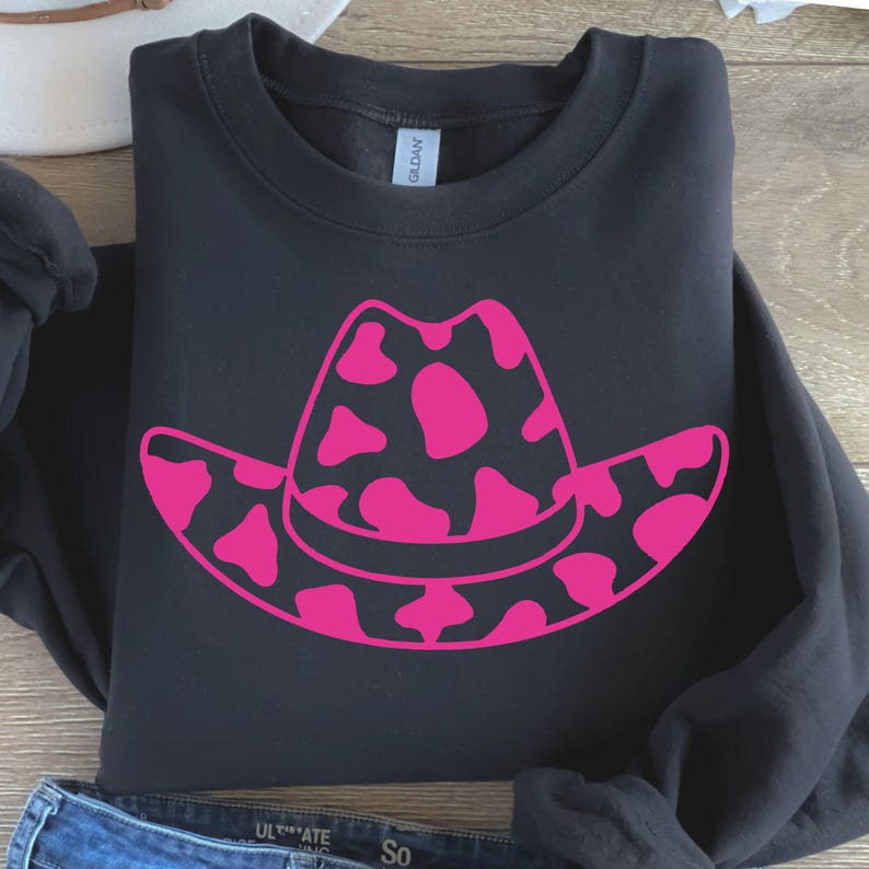 May include: Black sweatshirt with a large pink cowboy hat graphic, featuring a cow print design. The hat is centered on the front of the folded garment. The sweatshirt is made of a soft material.