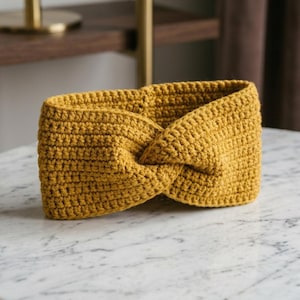 May include: Mustard yellow crochet headband with a twisted knot design. The handmade ear warmer is made of a thick, textured yarn and is displayed on a marble surface. This accessory is perfect for keeping ears warm during colder months.