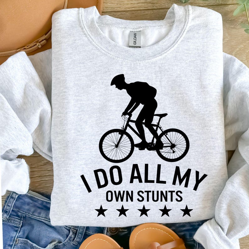 May include: A light gray sweatshirt with a black graphic of a cyclist performing a stunt. The text "I DO ALL MY OWN STUNTS" is below the graphic, with five black stars. The sweatshirt is made of a soft material.