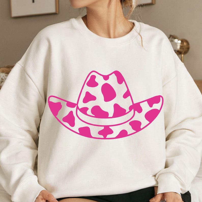 May include: A white sweatshirt with a pink cowboy hat design. The hat has a cow print pattern in pink. The sweatshirt is a casual style, suitable for everyday wear. The design is bold and eye-catching.