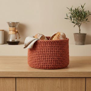 May include: A round, rust-colored crochet basket filled with two loaves of bread and a linen napkin. The basket sits on a wooden surface, with a coffee maker and a small potted plant in the background.