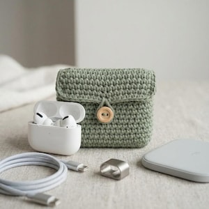 May include: A sage green, crocheted case with a wooden button closure, next to a white charging case with earbuds, a charging cable, a silver ring, and a wireless charger.