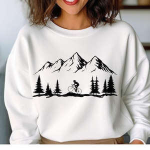 May include: White sweatshirt featuring a black mountain range, pine trees, and a cyclist. The graphic design is a silhouette style, creating a minimalist outdoor theme. The sweatshirt is a crew neck style.