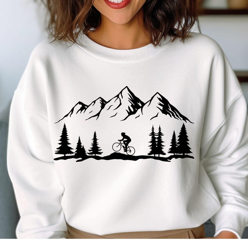 May include: White sweatshirt featuring a black mountain range, pine trees, and a cyclist. The graphic design is a silhouette style, creating a minimalist outdoor theme. The sweatshirt is a crew neck style.