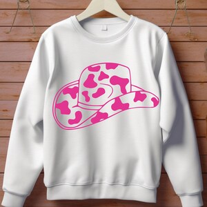 May include: White sweatshirt with a pink and white cow print cowboy hat design. The hat graphic is a striking feature on the long-sleeved top. The sweatshirt is displayed against a wooden backdrop.