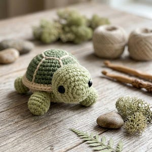 Op de afbeelding: Een kleine, handgemaakte gehaakte schildpad met een lichtgroene kop en ledematen, een donkergroen schild met crèmekleurige stiksels en twee zwarte plastic kraaloogjes. De schildpad is gemaakt met een getextureerde garenstructuur en een ronde vorm.