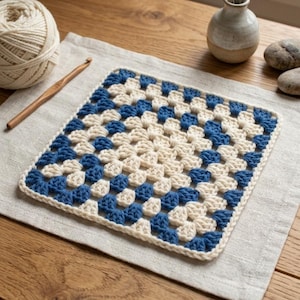 May include: A square crochet granny square featuring a repeating pattern of cream and deep blue yarn clusters. The square is worked in a classic cluster stitch design with a solid cream border edge, showcasing a balanced geometric colour arrangement.