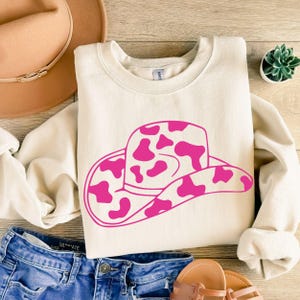 May include: Cream-coloured sweatshirt with a pink cowboy hat design featuring a cow print. The sweatshirt is on a wooden surface with blue denim shorts, a brown hat, and sandals.