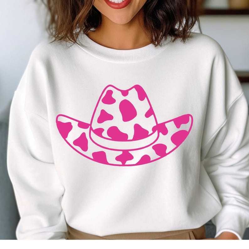 May include: A white sweatshirt with a pink cowboy hat graphic. The hat has a cow print pattern. The design is simple and bold, with the pink hat contrasting against the white fabric of the sweatshirt.