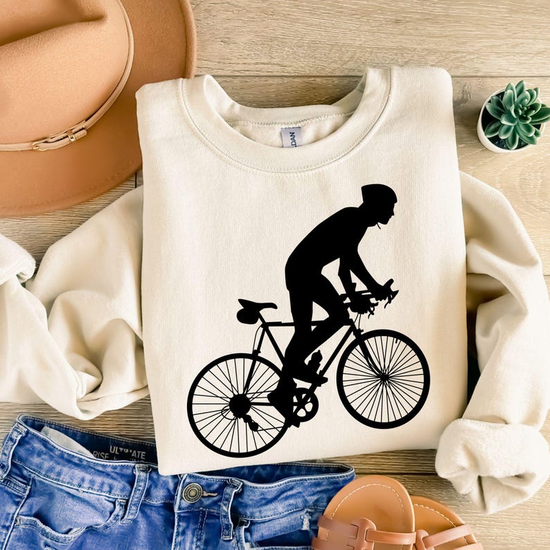 May include: Cream-colored sweatshirt featuring a black silhouette of a cyclist on a bicycle. The design is centered on the front of the sweatshirt. The image is on a wooden surface, with a hat, jeans, and sandals in the background.