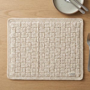 Könnte beinhalten: Cremefarbener rechteckiger Tischset mit gewebtem Design. Das Tischset hat eine strukturierte Oberfläche und einen genähten Rand. Ein kleines Etikett mit dem Text "LIVLIG HANDMADE" befindet sich in der unteren rechten Ecke. Ein Teller und Besteck sind im Hintergrund.