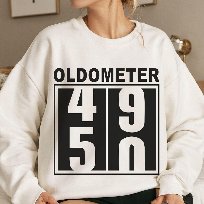 May include: Cream-colored sweatshirt with the word "OLDOMETER" above a black rectangle containing the numbers "49" and "50" in white. The sweatshirt is a casual, comfortable style.