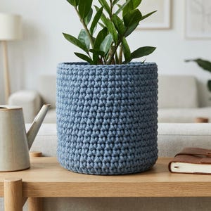 Könnte beinhalten: Ein blauer gehäkelter Blumentopf mit einer grünen Pflanze. Der Topf ist zylindrisch und steht auf einem hellen Holztisch. Eine Gießkanne und ein Notizbuch befinden sich ebenfalls auf dem Tisch. Der Hintergrund umfasst ein Sofa und eine Lampe.