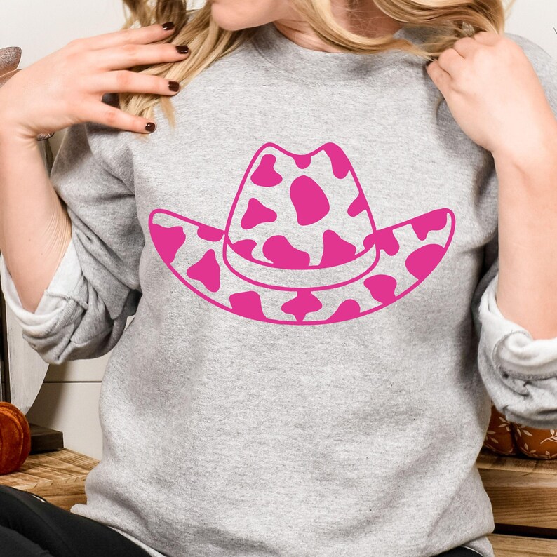 May include: A grey sweatshirt with a pink cowboy hat design. The hat has a spotted pattern. The sweatshirt is a casual style, suitable for everyday wear. The image shows a close-up of the sweatshirt.