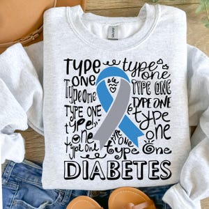 May include: A light gray sweatshirt with the words "Type One Diabetes" in a black, handwritten font. A blue and gray ribbon graphic is centered on the front. The sweatshirt is made of a soft material.